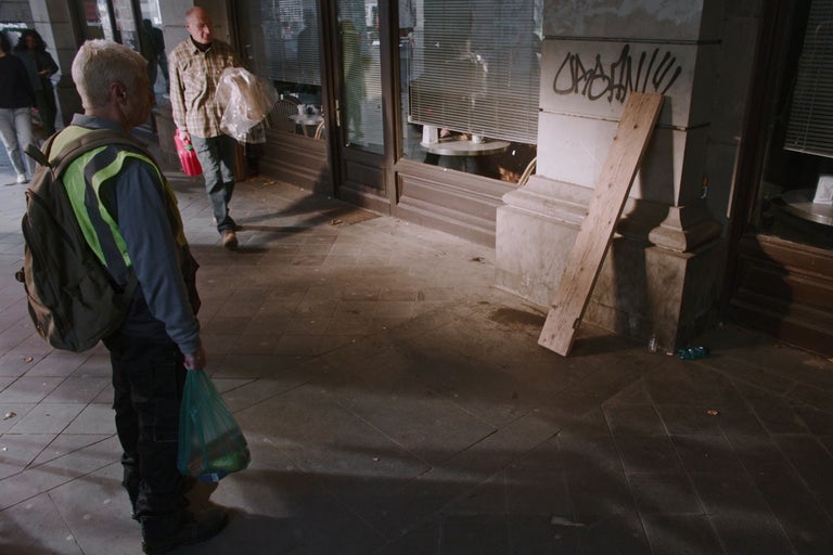 Scène urbaine extérieure avec une planche de bois contre une façade en pierre et un homme en gilet de sécurité au premier plan.