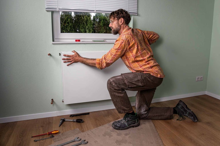 Un homme est agenouillé devant un mur et installe un radiateur. Des outils sont posés sur le sol.