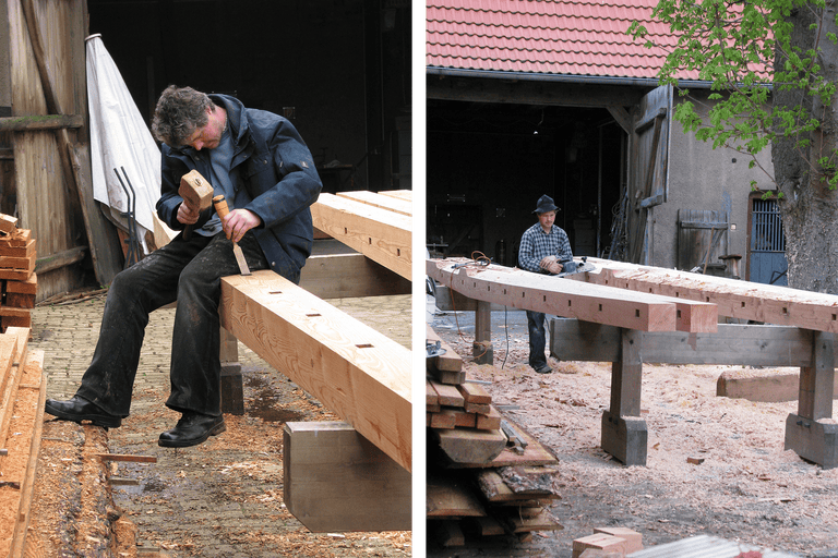 Deux artisans travaillent des poutres en bois avec des outils dans un atelier.