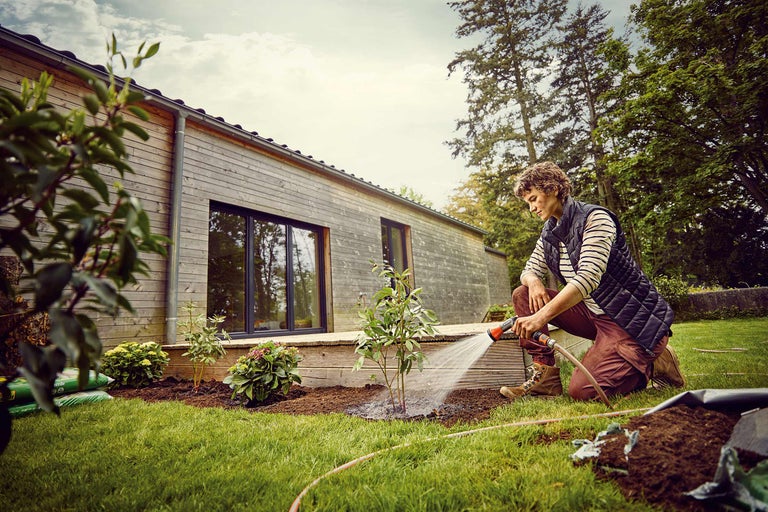 Une personne arrose de jeunes plantes dans un jardin avec une maison en arrière-plan.