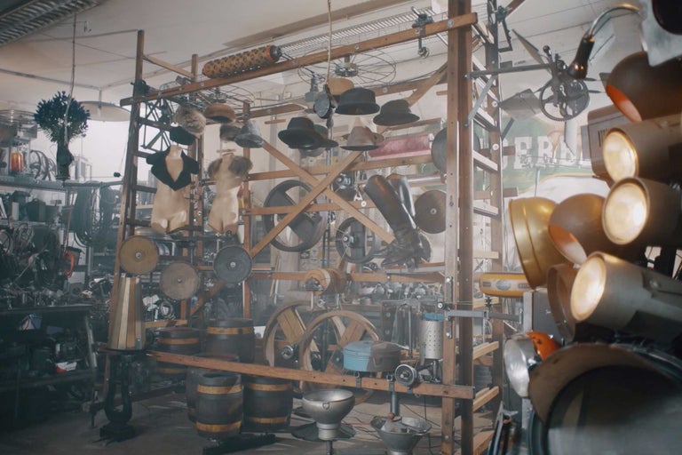 Szene mit Hüten, Kleidung, Fässern, Rädern und Scheinwerfern auf einer Holzkonstruktion in einem Raum.