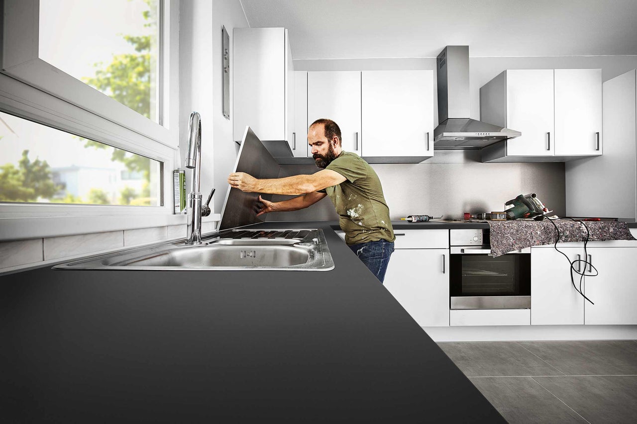 Un homme installe une crédence de cuisine dans une cuisine moderne avec évier, robinet et armoires blanches.