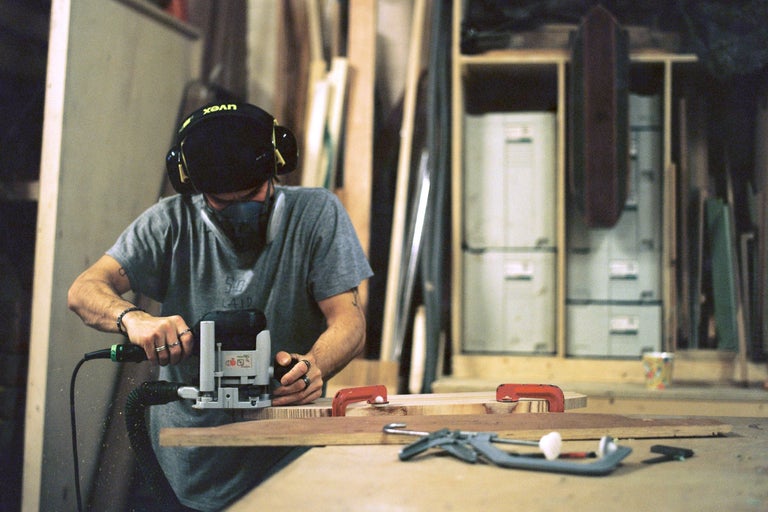 Un homme fraise du bois avec une défonceuse dans un atelier, avec des outils et des matériaux en arrière-plan.