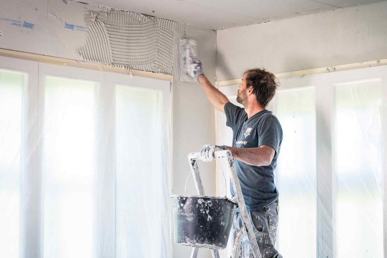 Un homme sur une échelle applique du plâtre au-dessus d'une fenêtre avec une truelle. Photo d'intérieur.