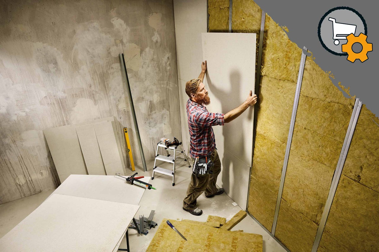 Un homme monte une plaque de plâtre sur un mur avec du matériau isolant et des profilés métalliques. D'autres plaques et outils sont posés au sol.
