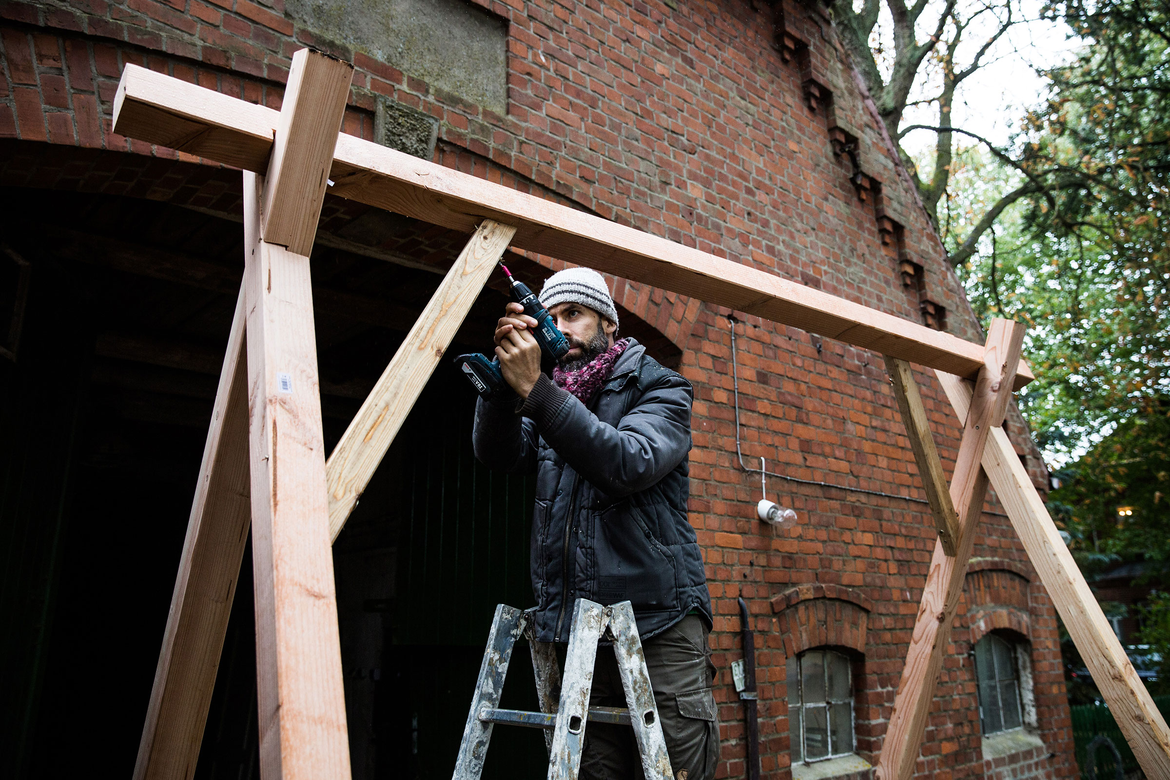 Un homme travaille avec une visseuse sans fil sur une construction en bois devant un bâtiment en briques.