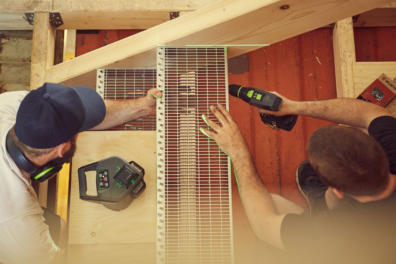 Deux artisans fixent une grille à une structure en bois à l'aide d'un niveau laser et d'une visseuse sans fil.
