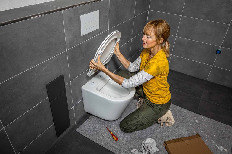 Une femme installe un siège de WC dans une salle de bains avec des carreaux gris et des toilettes suspendues.