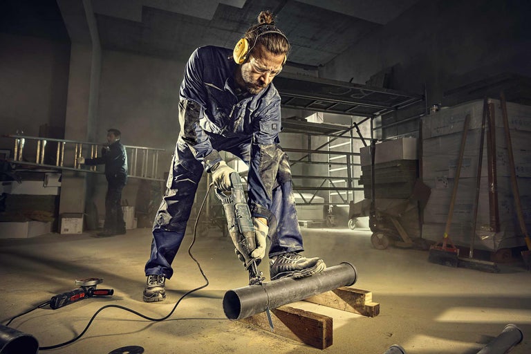 Un artisan équipé de protections de travail coupe un tuyau avec une scie sabre. À l'arrière-plan, des matériaux de construction sont entreposés.