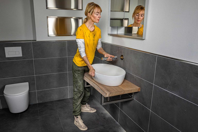 Salle de bains avec lavabo en céramique, plateau de table en bois et installation de robinetterie par une femme