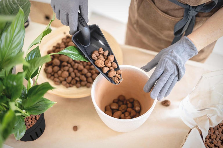 Une personne portant des gants de jardinage remplit un bol de billes d'argile à l'aide d'une pelle.