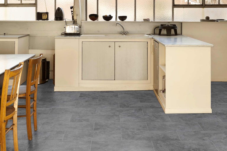 Küche mit grauem Bodenbelag in Fliesenoptik, Küchenzeile und Stühlen.