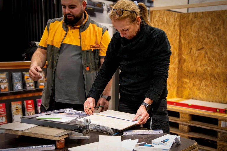 Zwei Personen vermessen eine Fliese mit einem Bleistift und einem Lineal auf einem Tisch mit Werkzeugen.