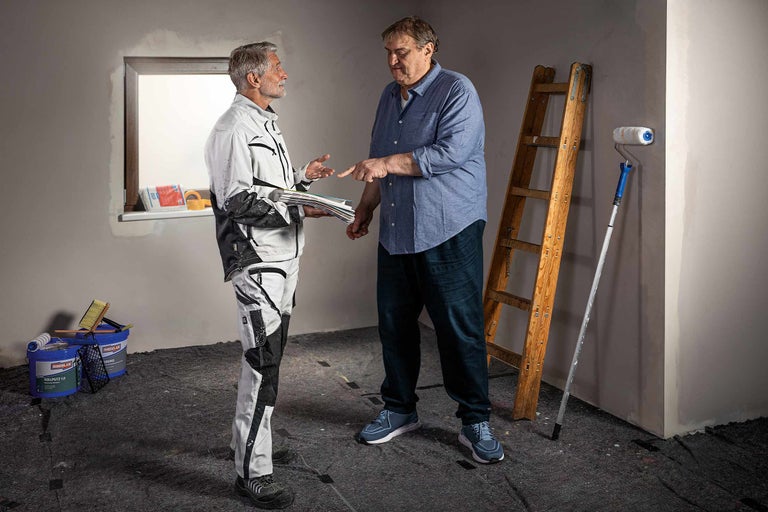 Zwei Personen besprechen eine Renovierung mit Leiter, Farbroller und Farbeimern in einem Raum.