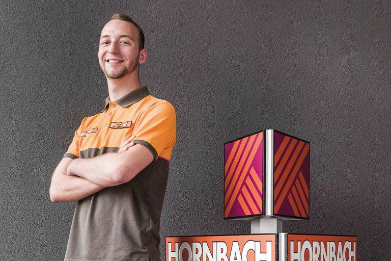 Ein lächelnder Hornbach Mitarbeiter neben einem Hornbach Leuchtschild