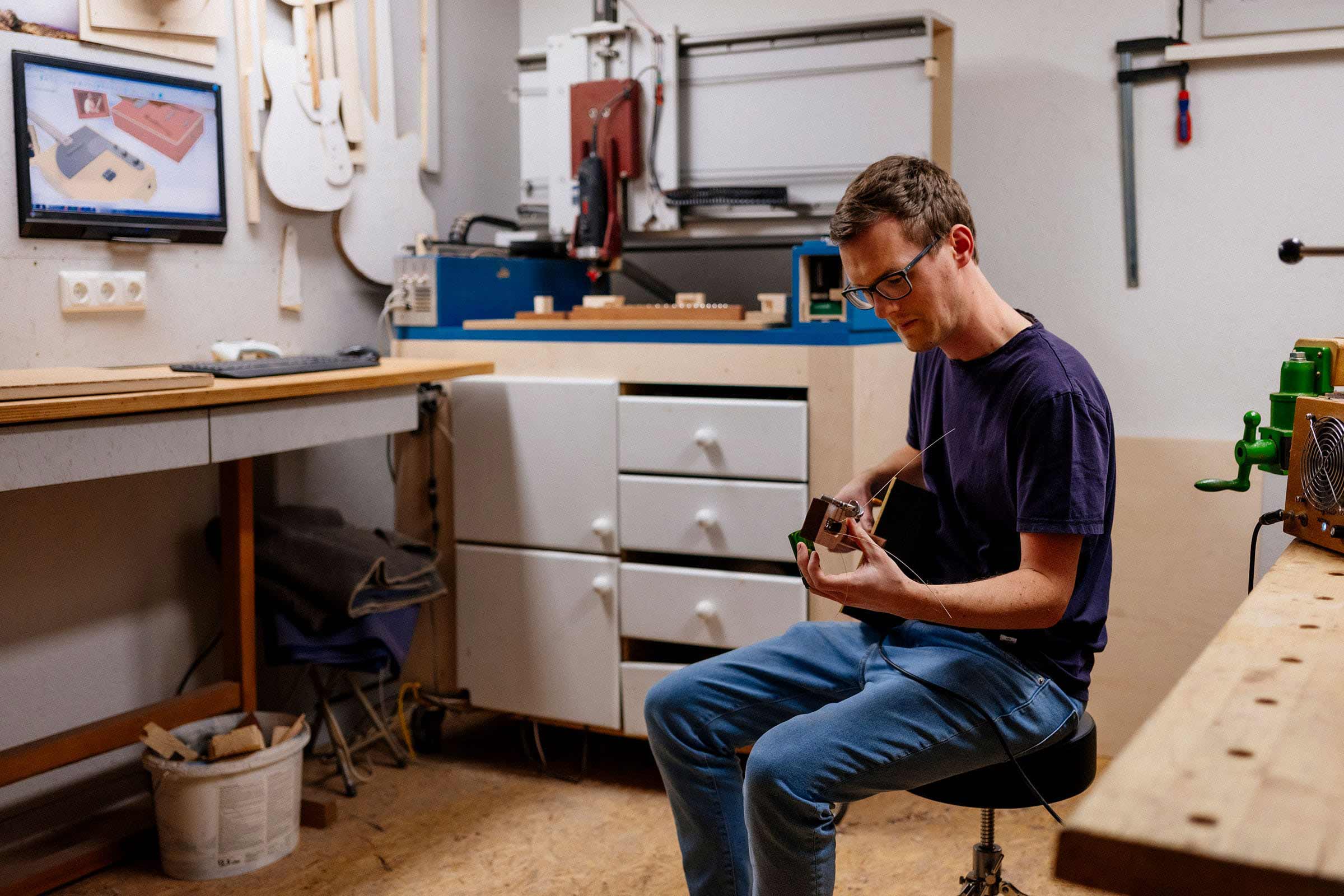 Ein Mann sitzt in einer Werkstatt und fertigt ein Musikinstrument, im Hintergrund sind eine CNC-Fräse, ein Bildschirm und eine Werkbank zu sehen.