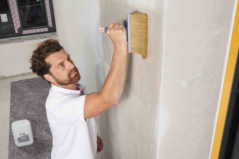 Un homme applique un apprêt en profondeur sur un mur à l'aide d'une brosse. Un châssis de fenêtre et un bidon d'apprêt en profondeur sont visibles en arrière-plan.