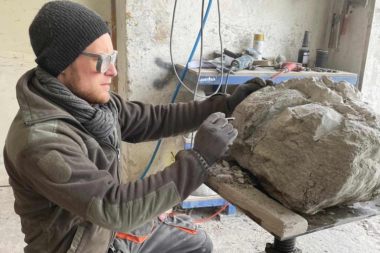 Ein Mann in Schutzkleidung bearbeitet einen Stein mit einem Werkzeug in einer Werkstatt.