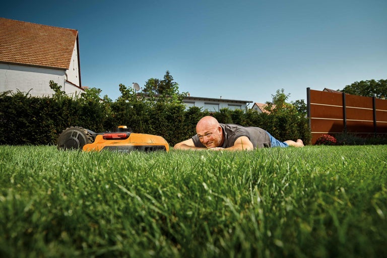 Mann liegt entspannt auf dem Rasen neben einem Rasenroboter