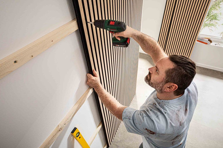 Un homme fixe un panneau mural acoustique à une latte en bois avec un tournevis sans fil.