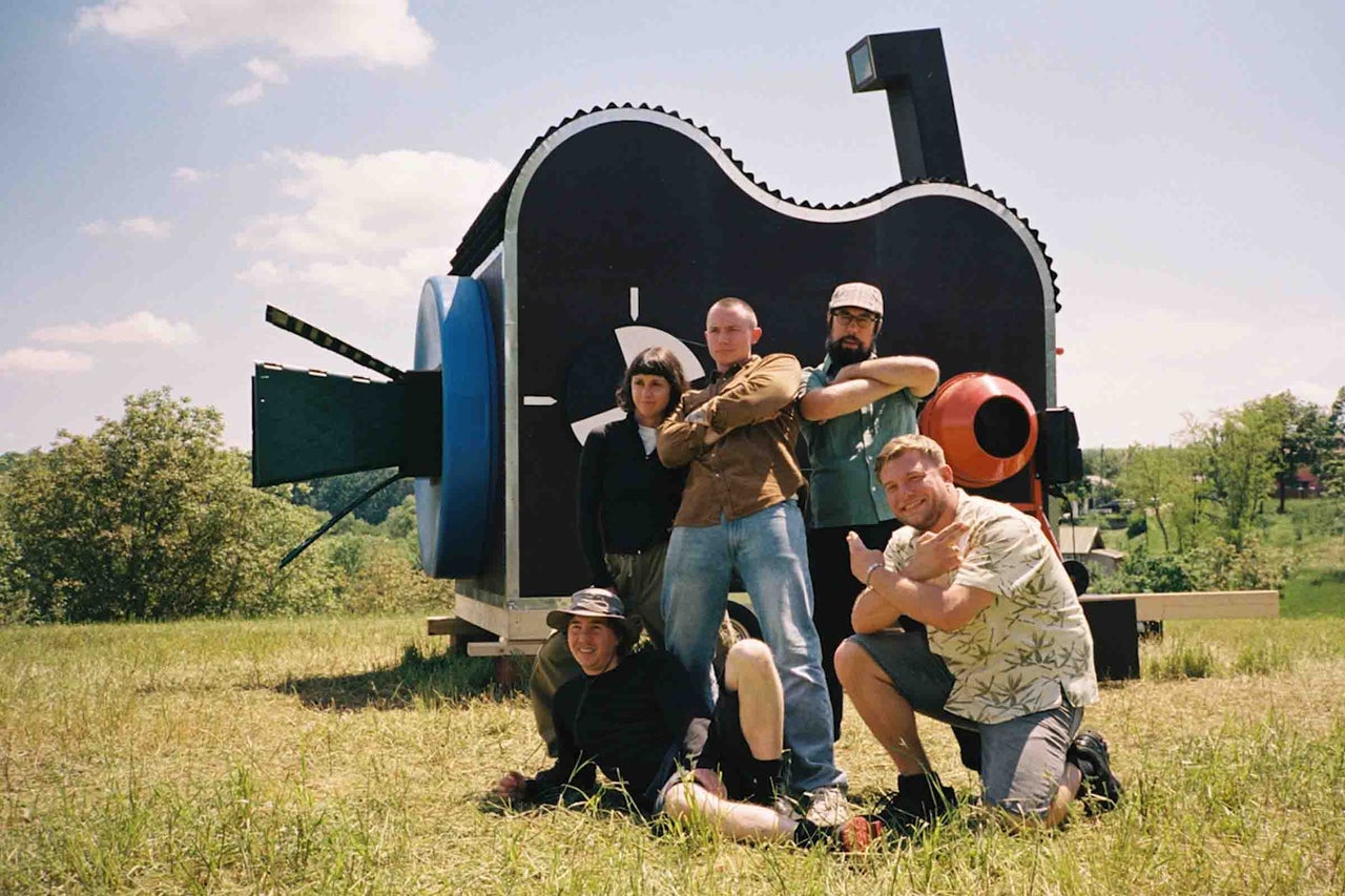 Un groupe de cinq personnes pose devant une grande construction en forme de caméra dans un pré.