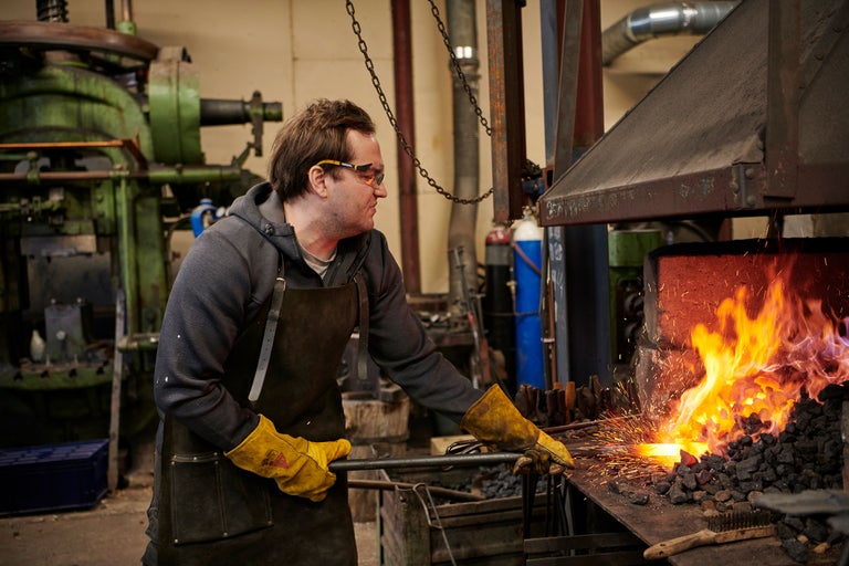 Ein Schmied mit Schutzbrille und Schürze bearbeitet glühendes Metall mit einer Zange in einer Schmiede.