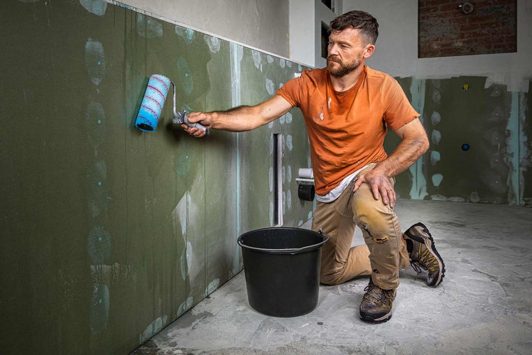 Artisan peint un mur avec un rouleau à peinture dans une pièce, seau posé au sol