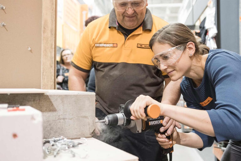 Eine Frau bohrt mit einer Bohrmaschine in ein Werkstück, während ein Mann zusieht, beide tragen eine Schutzbrille und die Frau eine Arbeitskleidung mit dem Hornbach Logo.