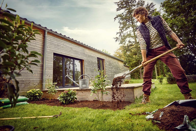 Un homme bêche la terre dans le jardin devant une maison à façade en bois.