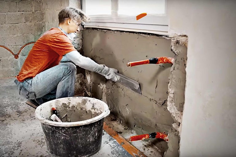 Un artisan applique du plâtre sur un mur, des tuyaux sont visibles.