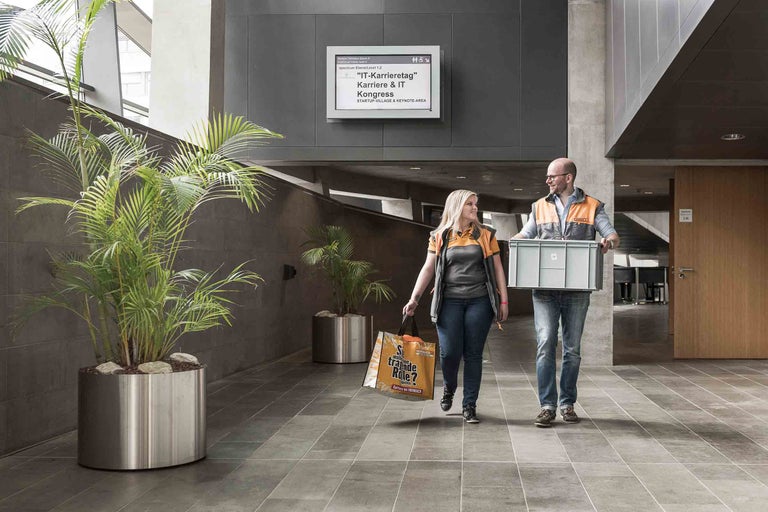 Ein Mann und eine Frau tragen eine Kiste und eine Einkaufstasche in einem Bürogebäude. Auf einer Anzeigetafel ist der IT-Karrieretag zu sehen.