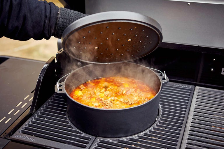 Un four hollandais avec couvercle est posé sur un barbecue, rempli d'un plat dont la vapeur s'échappe.