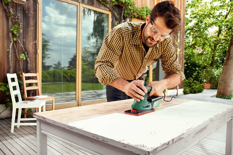Ein Mann schleift eine Tischplatte mit einem Schleifgerät im Freien.