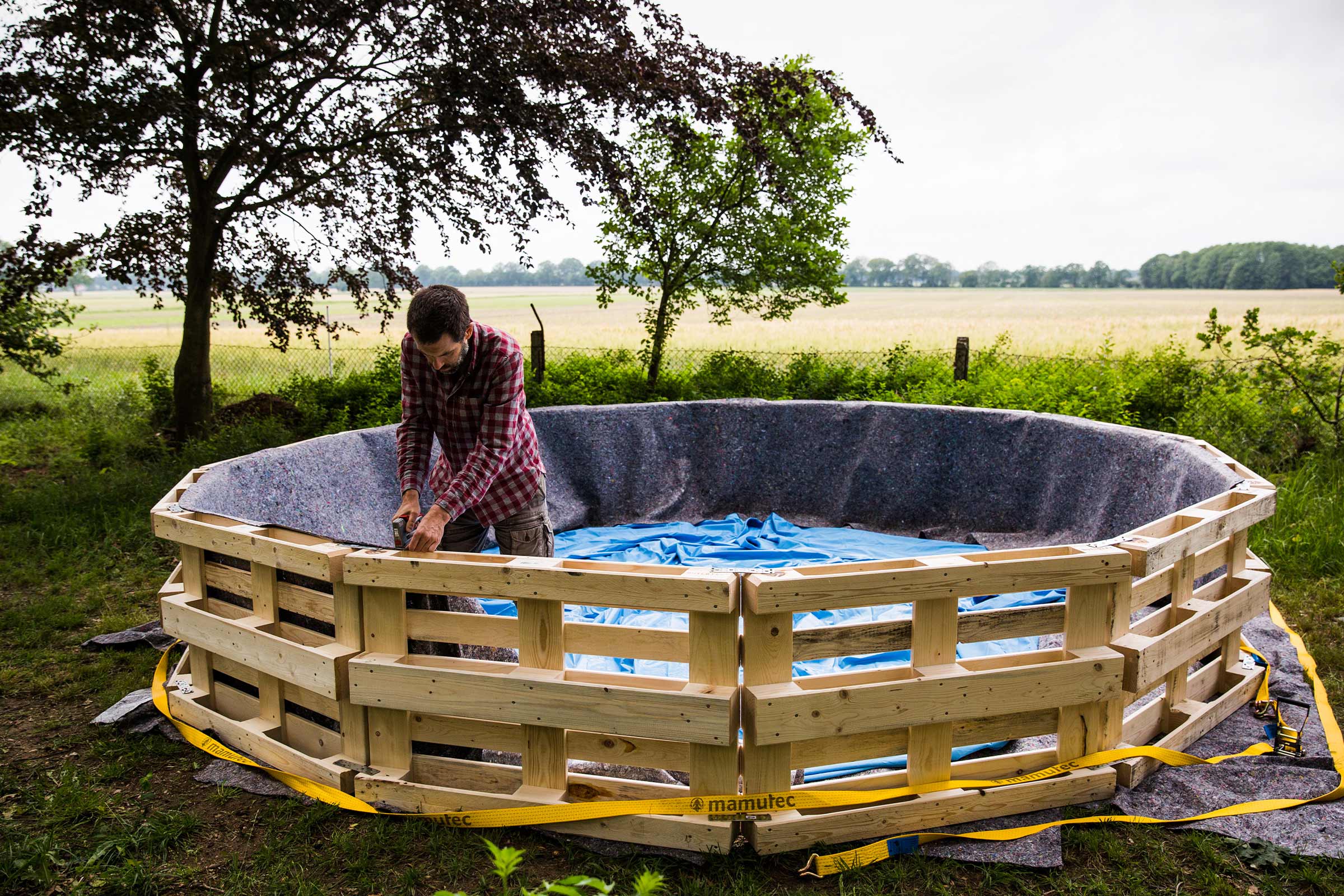 Un homme construit une piscine en palettes dans le jardin, recouverte de film et de non-tissé