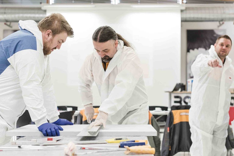 Drei Handwerker in Schutzanzügen bearbeiten eine weiße Oberfläche mit Werkzeugen