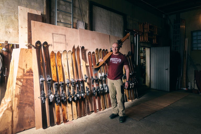 Mann mit Holzski vor Holzwand mit Ski-Kollektion