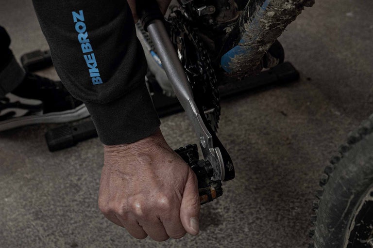 Eine Person benutzt einen Schraubenschlüssel an einem Fahrradpedal, im Hintergrund ist ein Bike Broz Pullover zu sehen.