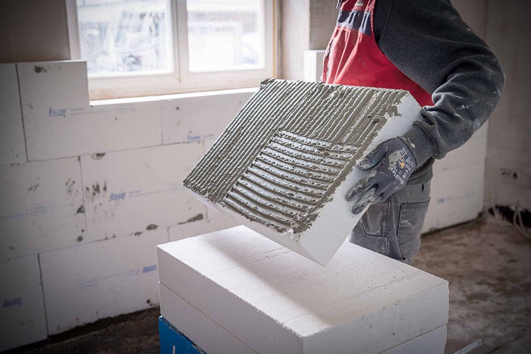 Ein Bauarbeiter hält einen Porenbetonstein mit aufgetragenem Kleber in den Händen, um eine Wand zu bauen.