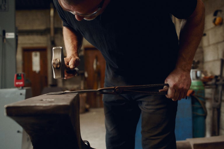 Ein Schmied bearbeitet ein Metallstück mit einem Hammer auf einem Amboss.