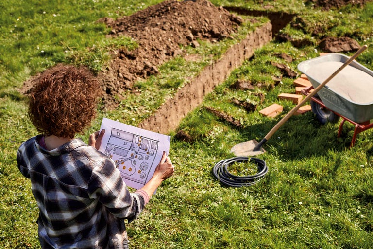 Femme regarde un plan de construction dans un jardin avec pelle, brouette, tuyau et tranchée creusée