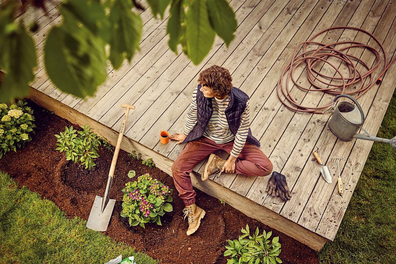 Scène avec personne sur terrasse en bois, outils de jardinage, arrosoir, tuyau d'arrosage et plantes