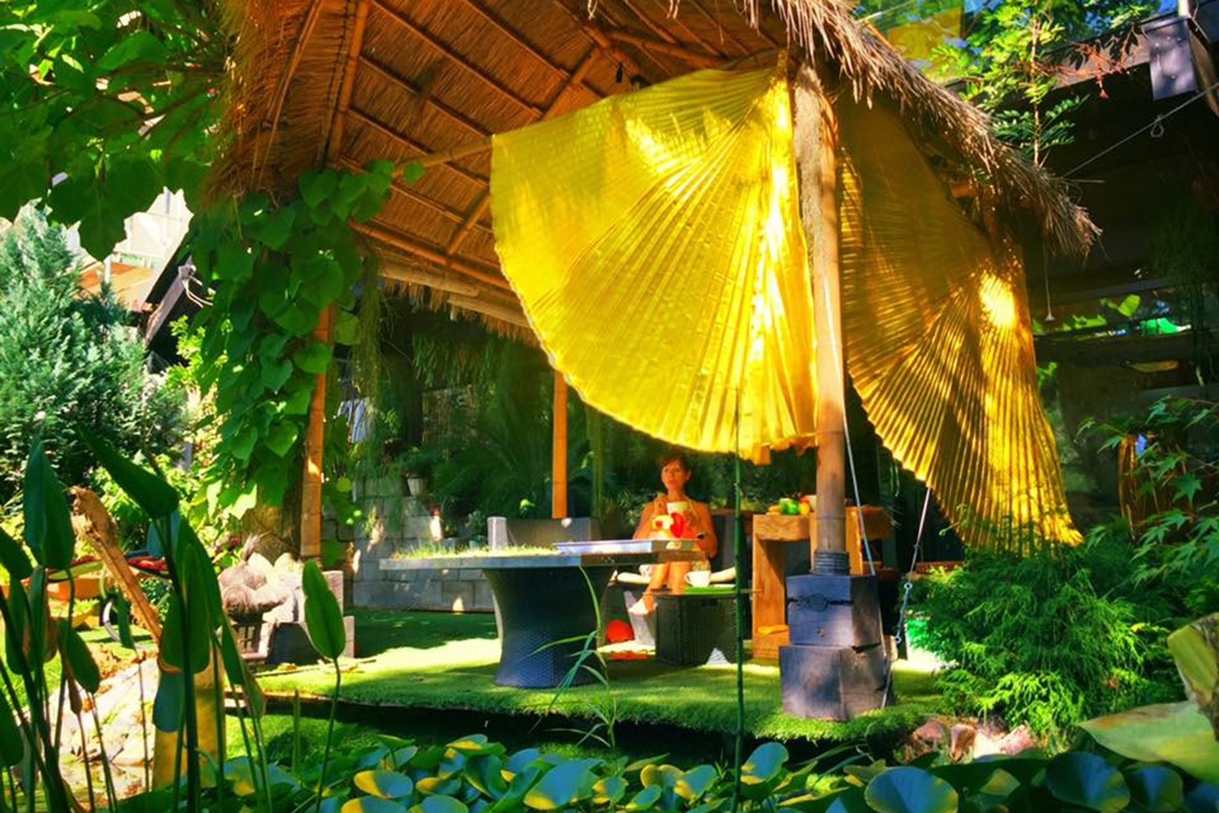 Gartenpavillon mit Strohdach, Tisch und Stühlen in üppiger grüner Umgebung
