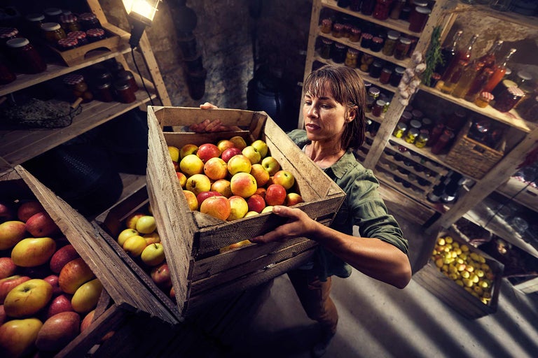 Une femme porte une caisse en bois pleine de pommes dans un entrepôt avec des étagères remplies de provisions.