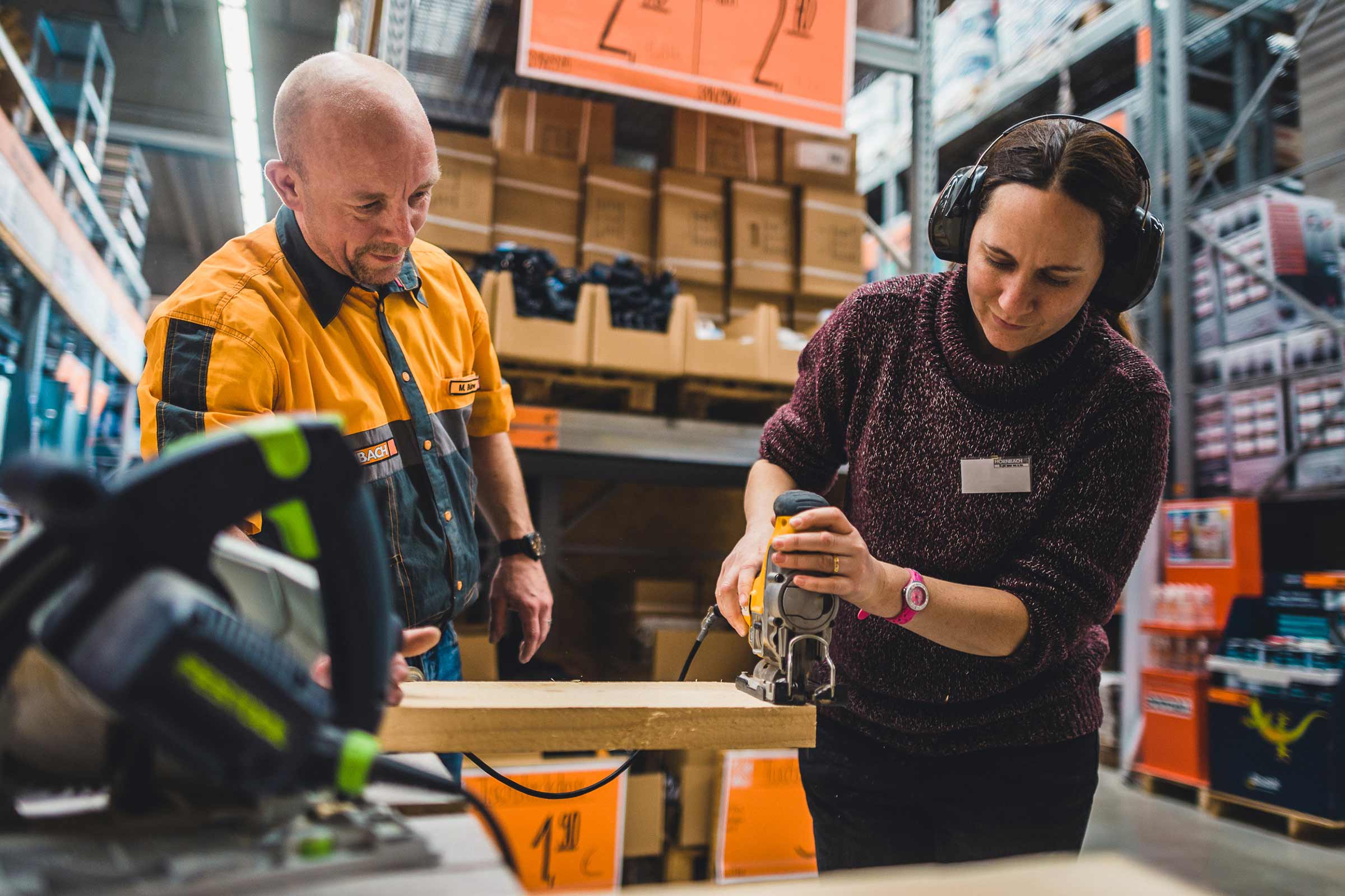 Une femme portant des protections auditives scie une planche de bois avec une scie sauteuse pendant qu'un homme regarde.