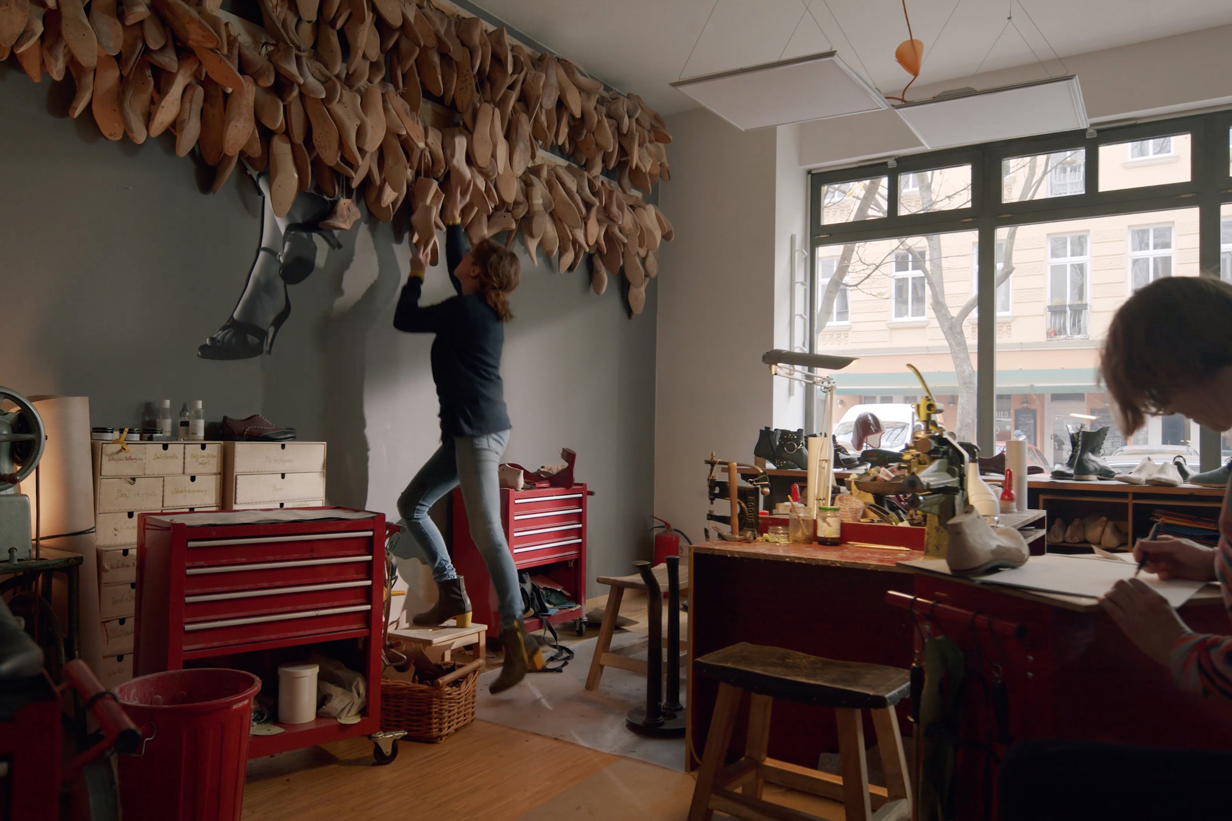 Innenansicht eines Schuhgeschäfts mit Werkzeugen, Werkbänken und einer Person, die an der Wand Holzschuhleisten anbringt.