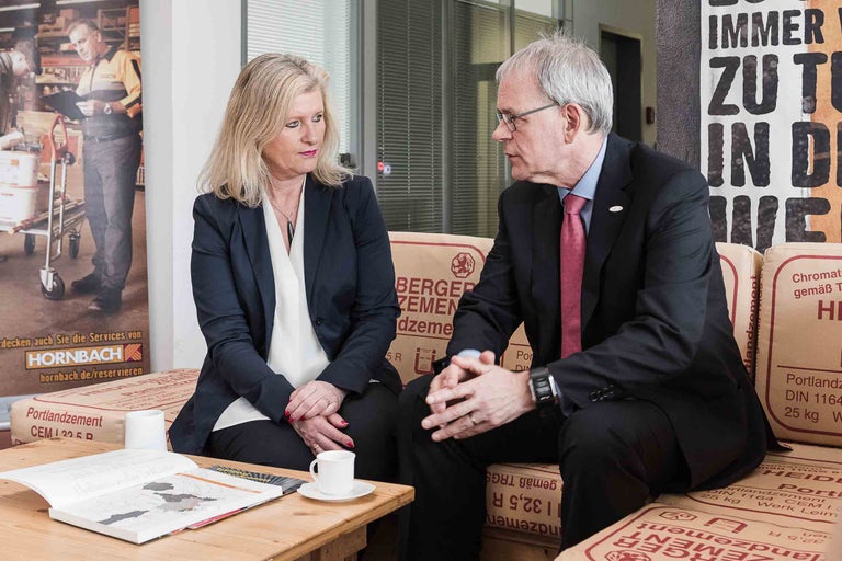 Zwei Personen sitzen auf Zementsäcken an einem Tisch mit Broschüre und Kaffeetasse.
