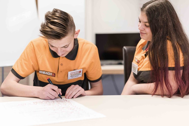 Zwei Hornbach Mitarbeiter sitzen an einem Tisch. Einer der beiden schreibt etwas auf ein Blatt Papier.