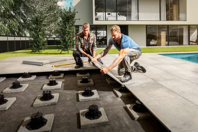 Deux artisans posent des dalles de terrasse sur des plots dans un jardin.