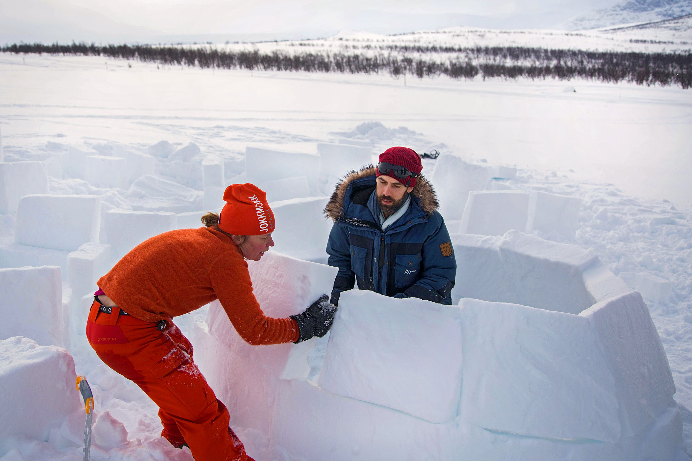 Zwei Personen bauen ein Iglu aus Schnee in einer Winterlandschaft.