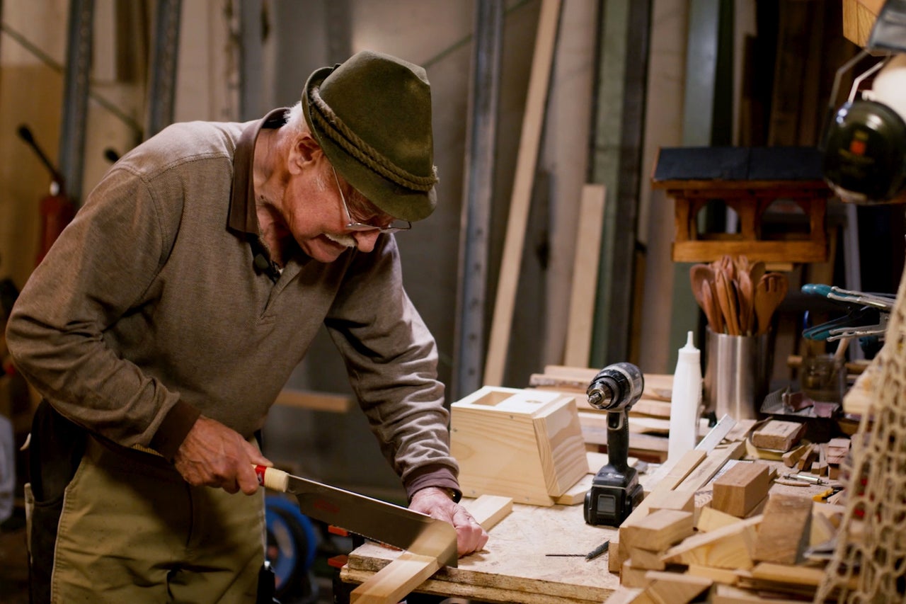 Un homme scie du bois dans un atelier avec une scie à main, entouré d'outils et de matériaux.